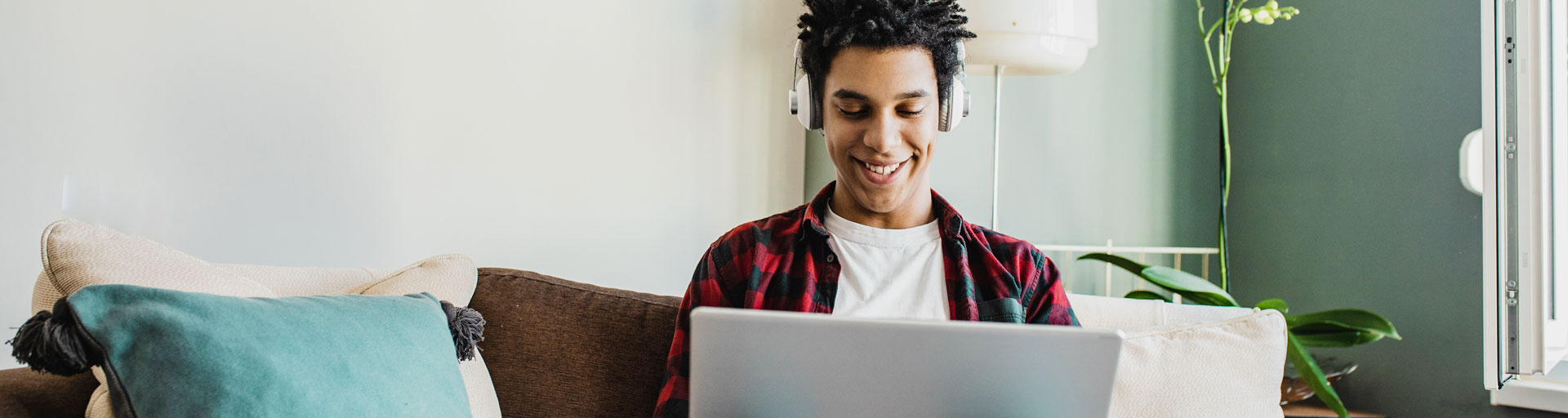 male student sitting on a couch, wearing headphones smiling at the laptop he is using